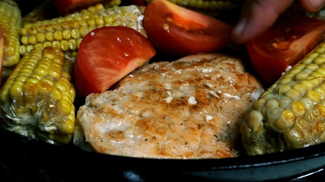 Delicious home cooking. Young cobs of corn and chicken fillet steak are fried in a pan and add chopped tomatoes. Close-up.