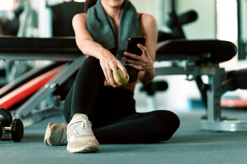 A girl hold green apple using smartphone in fitness gym. Woman exercise in gym fitness breaking and relax.