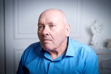Obraz premium Portrait of an elderly bald man in a blue shirt in the room. A poor pensioner lives alone.
