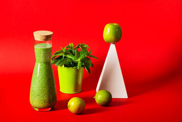 A beautiful bottle with a green smoothie stands on a red background, next to it stands a white geometric pyramid with a green apple, a bucket with mint leaves, and limes.