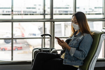 Young asian woman wearing sergical mask using smart phone while waiting for her flight at the airport