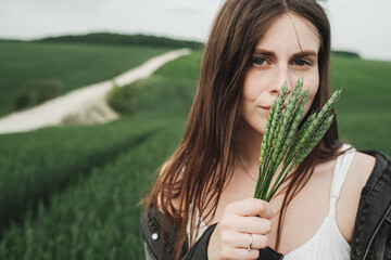 Young Stylish Girl in White Dress and Denim Jacket Holding Spikelets of Green Wheat in the Field