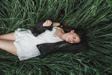 Young Stylish Girl in White Dress and Denim Jacket Lies in the Green Wheat Field