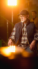 Protrait of Prominent Successful Senior Director Sitting in a Chair on a Break. On the Studio Film Set with High End Equipment Professional Crew Shooting High Budget Movie