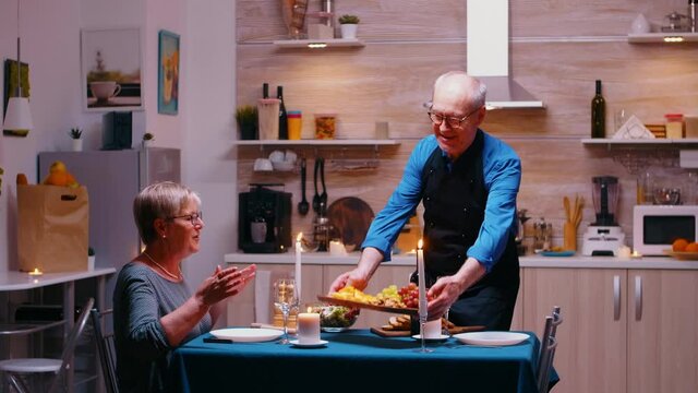 Retirement mature man surving dinner at a dining room. Senior old couple talking, sitting at the table in kitchen, enjoying the meal, celebrating their anniversary with healty food.