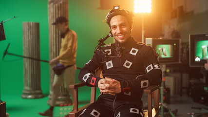 Handsome Smiling Actor Wearing Motion Capture Suit and Head Rig having Lunch Break, Sitting on Chair, Looking at Camera. Studio High Budget Movie. On Film Studio Period Costume Drama Film Set