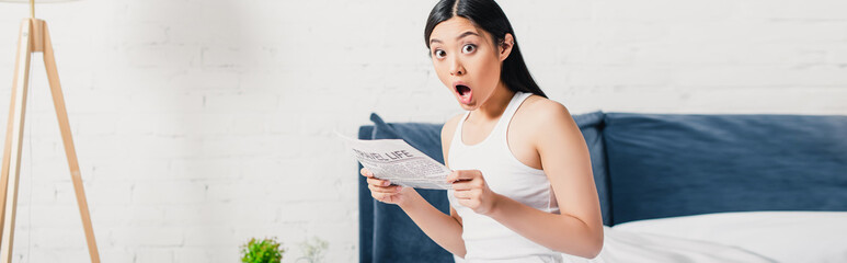 Horizontal crop of shocked asian woman holding newspaper on bed