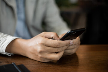 Fototapeta premium Close up of businessman's hands holding and typing on black cellphone for business communication in the cafe with a cup of coffe on wooden table. Business stock photo.