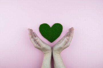 Earth day, ecology and Environmental preservation concept. Hands holding green heart from grass on pink background. Love nature and save the planet and world.