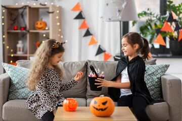halloween, holiday and childhood concept - smiling little girls in costumes with candies at home
