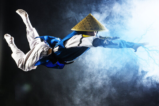 Cosplay Shaolin monk in a straw hat flies through the air releases lightning from his hands on a black background and with smoke