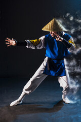 Cosplay Shaolin monk in a straw hat stands in a fighting stance hand extended forward with the effect of scattering in the air on a black background and with smoke