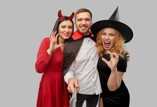 friendship, holiday and people concept - group of happy smiling friends in halloween costumes of witch, devil and vampire taking picture by selfie stick over grey background