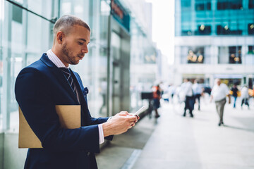  Confident male lawyer standing outdoors in business district of city dialling number on mobile phone, serious male entrepreneur checking email on smartphone outdoors sending feedback to colleague.