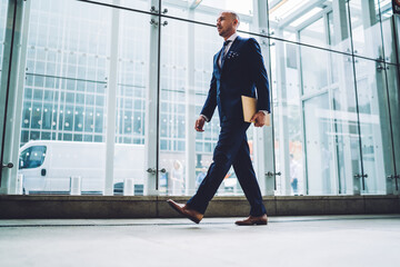 Fototapeta na wymiar Handsome male entrepreneur in elegant suit passing glass window of modern architecture building with copy space for advertising, confident financial manager walking with documents in business center .
