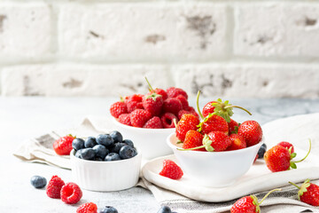Ripe berries. Different berries in white ceramic bowls on a light table. Strawberries, raspberries and blueberries in plates against a white brick wall. Space for text