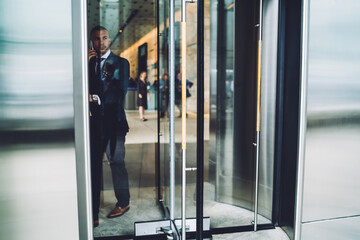 Serious executive manager leaving office building talking on mobile about business, male entrepreneur in elegant formal suit having phone conversation while going outdoors from modern estate.