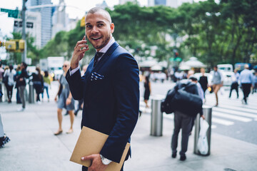 Cheerful businessman in formalwear talking on phone walking on street getting to work, successful financial manager excited with good news during mobile conversation outdoors in megalopolis.