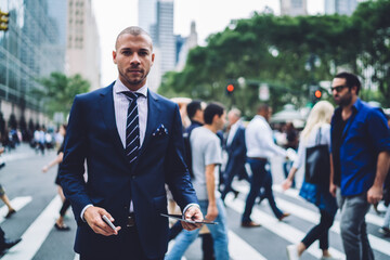 Portrait of serious male entrepreneur standing on crosswalk in new york city looking at camera, confident businessman dressed in elegant suit using modern technology while walking in office . - obrazy, fototapety, plakaty