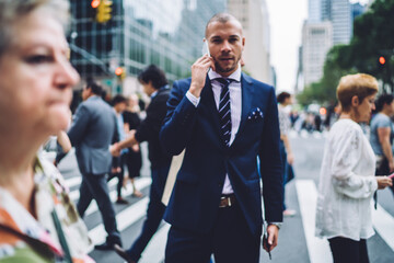 Handsome serious businessman in elegant wear making phone call in roaming using cheap tariffs while standing in crowded crosswalk, portrait executive manager having mobile talk on avenue of big city.