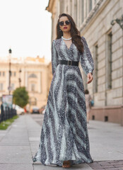 Naklejka premium Pretty female model in gray dress with stripes and sunglasses standing and posing at city street on a summer day. Full length fashion oudoor portrait