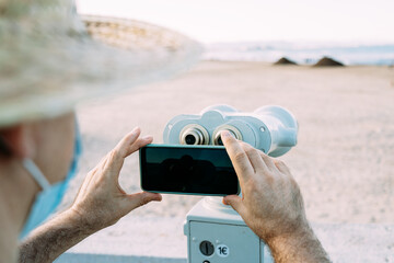 Close-up of a tourist wearing a hat and face mask taking pictures with binoculars at a viewpoint on the beach. 1€