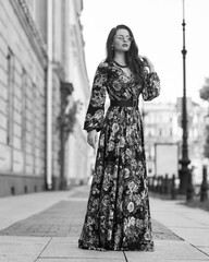 Young elegant lady in red and black floral design dress. Full length woman portrait. Beautiful girl standing and posing at city street. Fashionable female model with wavy hair