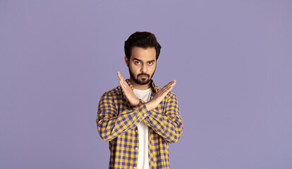 Handsome young Indian guy with crossed hands showing stop gesture over violet background