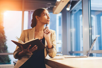 Portrait of skillful afro american female executive thinking over working schedule for employee writing report into notebook while using laptop computer and wireless connection in coworking space