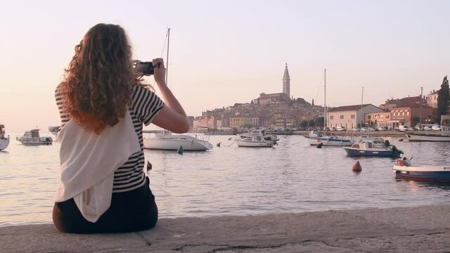 Young woman taking a picture of Rovinj marina and city view with boats and St. Euphemia cathedral at sunset. Istria, Croatia.
