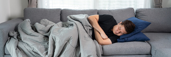 Sick and unwell Asian men holding his head, having a strong headache and sleeping on sofa at home wrapped in grey blanket