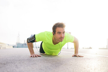 Handsome sports man with running armband doing push-ups on asphalt city road while listening to music with headphones,caucasian male jogger warm up before start his workout training outside in morning