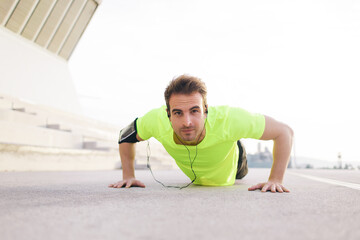 Portrait of strong build sports man doing push-ups on asphalt city road and listen to music with headphones, young male jogger warm up before start his workout training outside and look to the camera