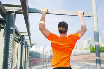 Back view of purposeful sports man with strong body engaged in active sports while pull up on the horizontal bar outdoors, young male runner in bright t-shirt training hard in urban setting at sunset