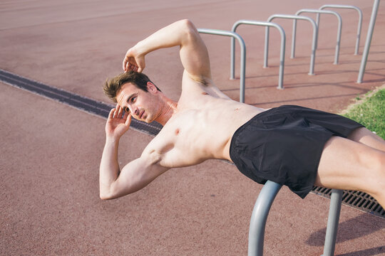 Half Length Portrait Of Handsome Sportive Man Doing Press Abdominal Exercise Outdoors In Summer Sunny Day, Muscular Build Male Runner Warming Up Before Began His Morning Run