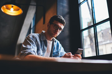 Portrait of confident hipster guy in optical eyeglasses looking at camera while chatting online in social networks on mobile phone sitting in university interior.Copy space area for advertising