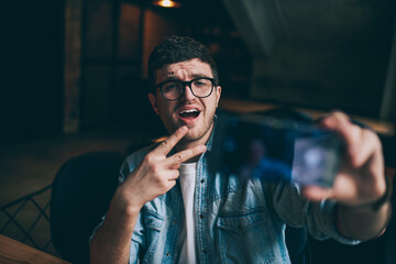 Excited male blogger showing peace fingers and taking funny pictures on mobile camera indoors, young emotional hipster guy making photos for social networks connected to wifi on smartphone device
