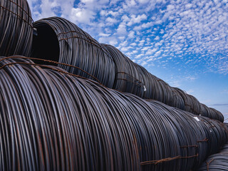 A hot rolled metal bar wound into rolls in a steel product warehouse under a blue sky with white clouds.