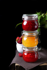 Raspberry, orange and strawberry jams in glass jars against the black background. Three jars of jam on the edge of the table
