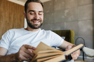 Image of young happy man smiling and reading book while sitting on couch