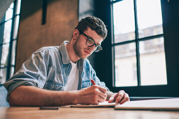 Serious male student writing essay sitting at desktop and spending time for course work, concentrated hipster guy doing school homework and planning study process, concept of education and knowledge