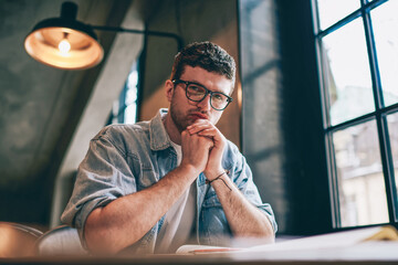 Portrait of serious hipster guy sitting at table and looking at camera indoors, concentrated man...