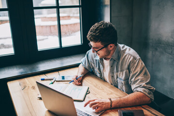 Hipster guy pondering on idea for essay sitting at wooden table with laptop device and looking on textbook, smart male freelancer spending time for distance job thinking about business indoors - obrazy, fototapety, plakaty