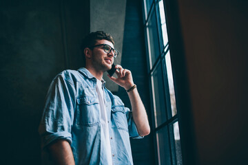 Positive male user in spectacles calling to friend via application on cellular phone connected to wifi indoors, casual dressed smiling hipster guy looking in window during smartphone conversation