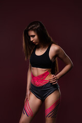 Kinesiology taping. Young female athlete on red background with kinesiology tape on tummy and hips. Fat lose, cellulite removal, sport physical therapy,recovery concept.