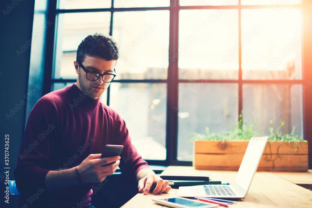 Poster male student in eyeglasses reading notification on smartphone while sitting in sunshine coworking sp