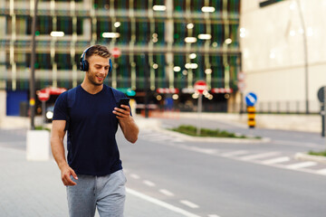 Young sport man walking in the city and listens to music via smartphone through wireless headphones