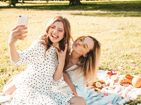 Two young beautiful hipster woman in trendy summer sundress and hats. Carefree women making picnic outside. Positive models sitting on plaid on grass, eating fruits and cheese Taking selfie photos
