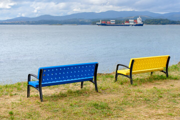 Naklejka na ściany i meble blue and yellow benches on the shore with a cargo ship on the horizon
