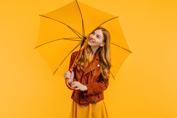 Pensive girl in a yellow dress and autumn jacket, with a yellow umbrella on an isolated yellow background